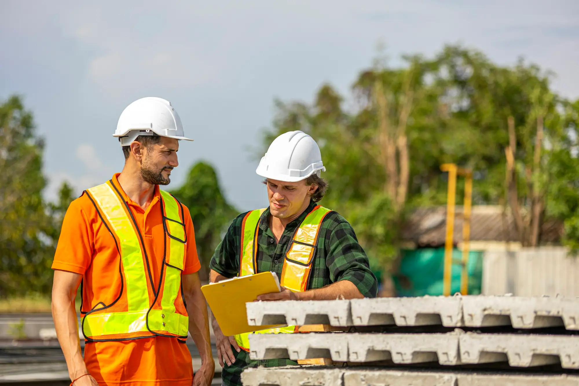 Two engineers in white hard hats and hi-vis vests reviewing a clipboard beside stacked precast concrete panels on an outdoor site