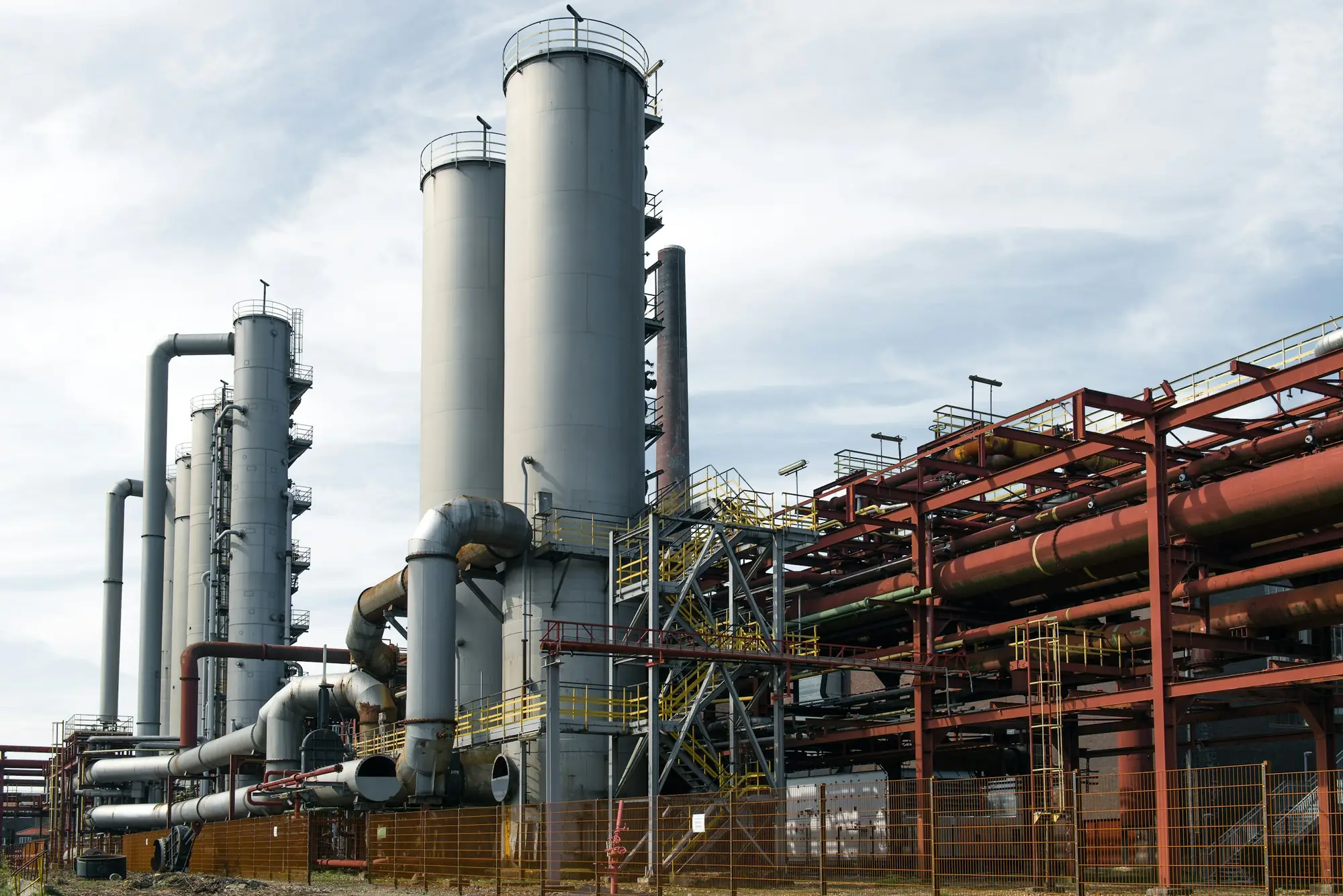 Tall grey process columns and ducting beside red steel pipe racks at a chemical processing plant on a bright overcast day